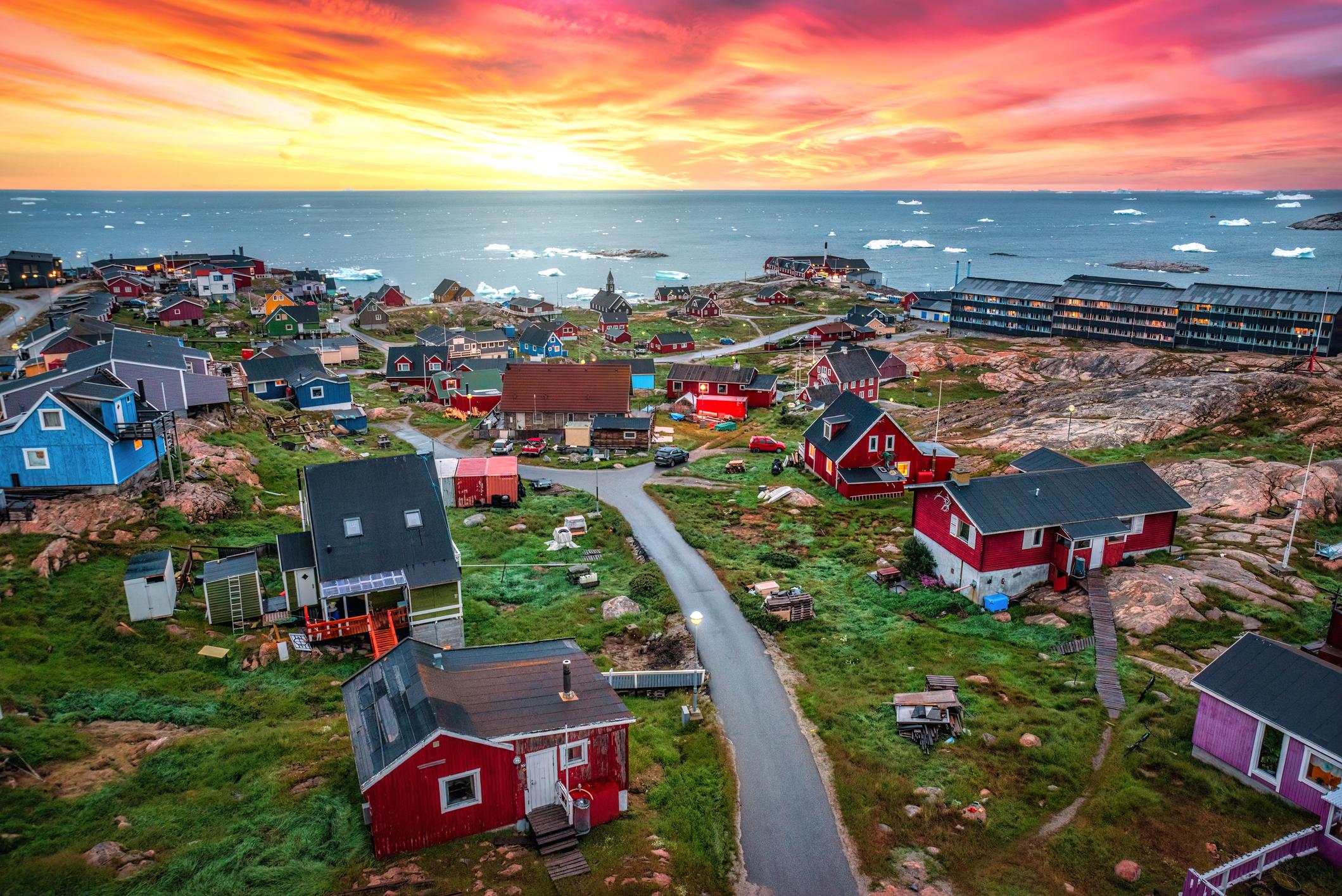 View of the Ilulissat village in Greenland featuring a colorful settlement with houses built on rugged terrain under a vibrant Arctic sunset, icebergs floating in the distant ocean. Midnight sun - Greenland eSim